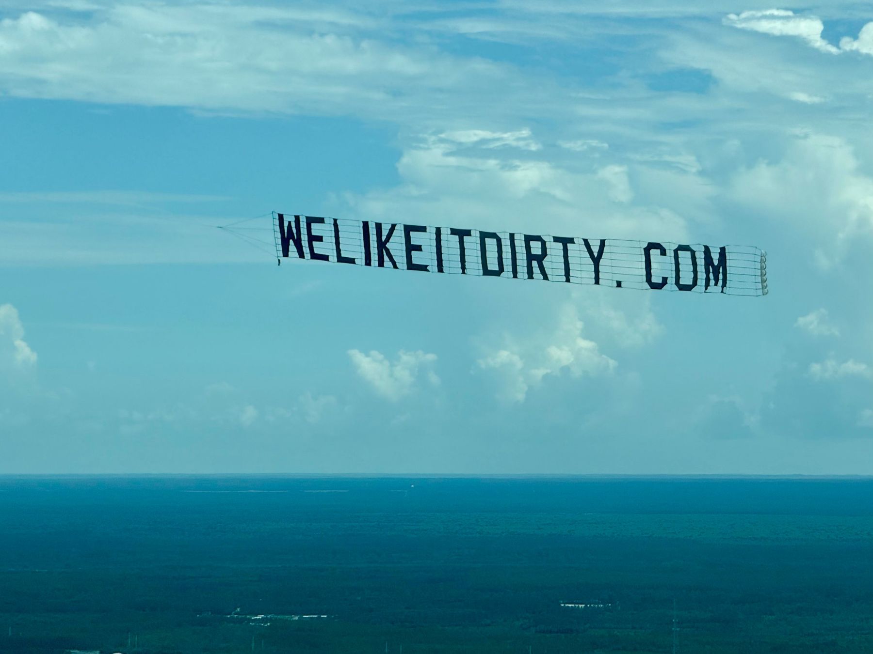 Letter banner towed across a blue sky above green countryside